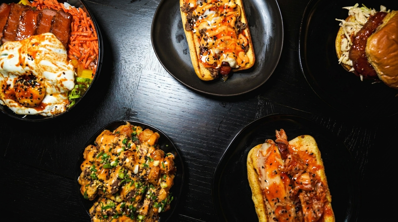 Overhead view of five dishes: a rice bowl, two hot dogs, a burger, and a sauced chicken dish.