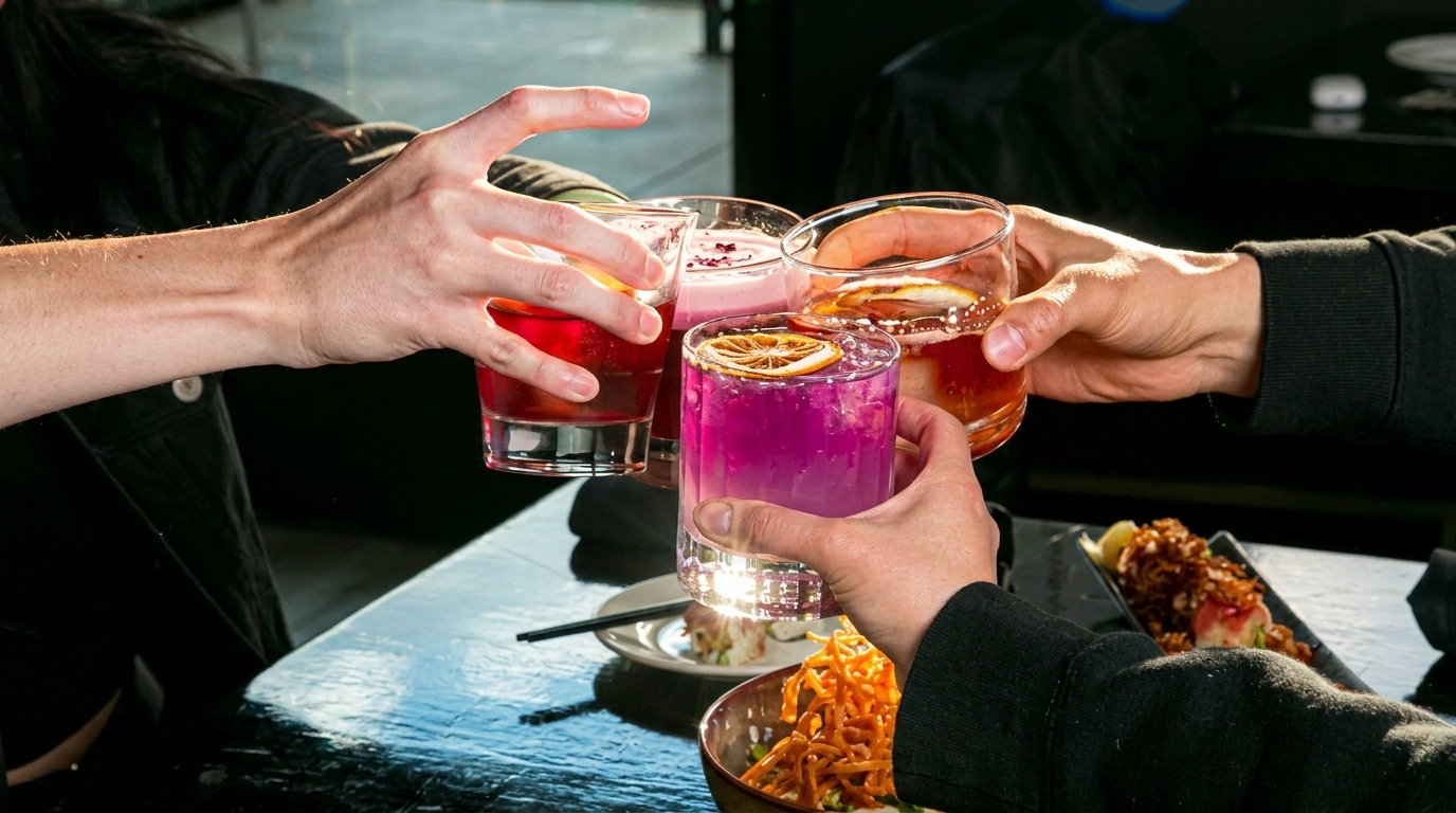 Four hands clinking colorful cocktails over a table with food, celebrating at a restaurant.
