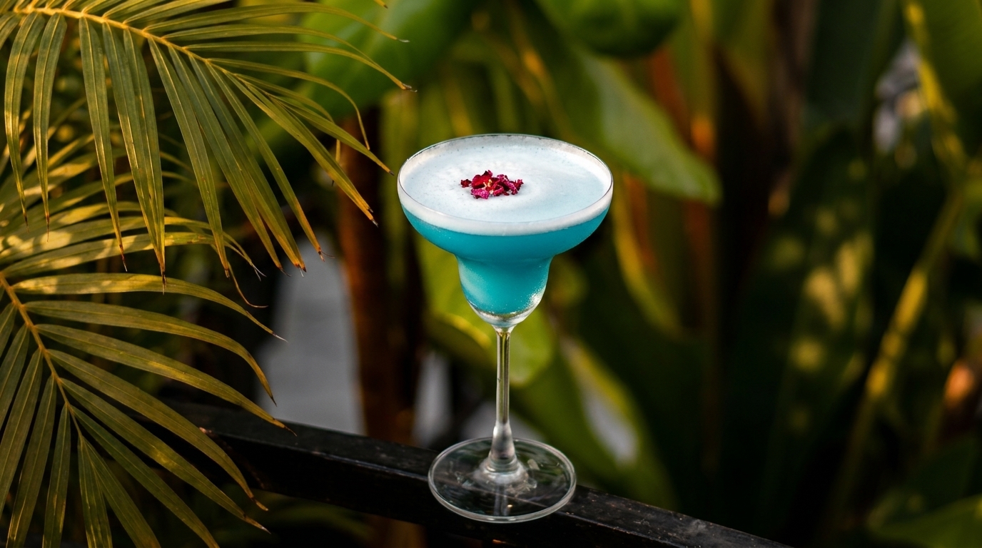 Vibrant blue cocktail with white foam and rose petals, set against lush green foliage.