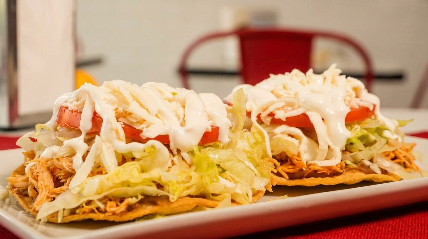 Two tostadas topped with shredded chicken, lettuce, tomato, cheese, and crema on a white plate.