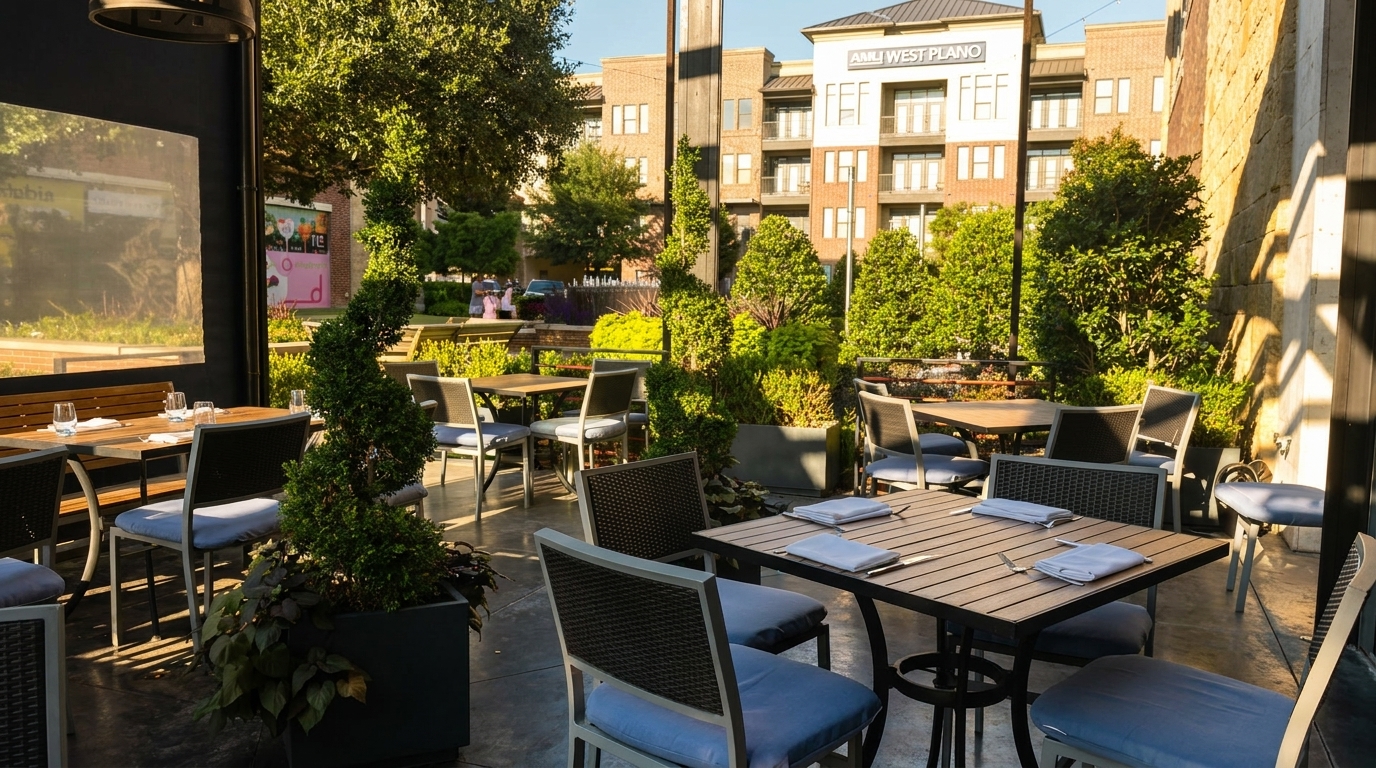 Outdoor patio dining area with tables, chairs, lush greenery, and a building in the background.