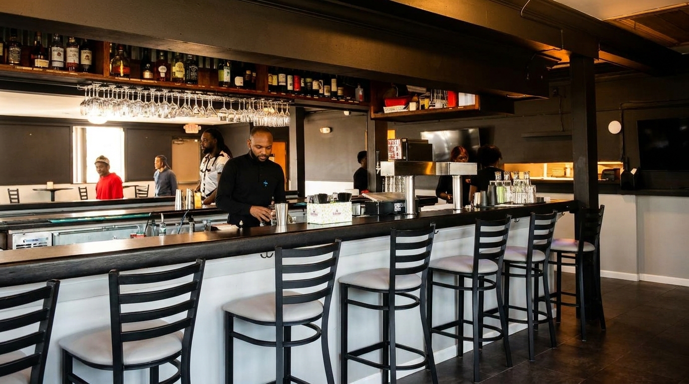 A bartender mixes drinks behind a long bar with stools, bottles, and glasses in a dimly lit restaurant.