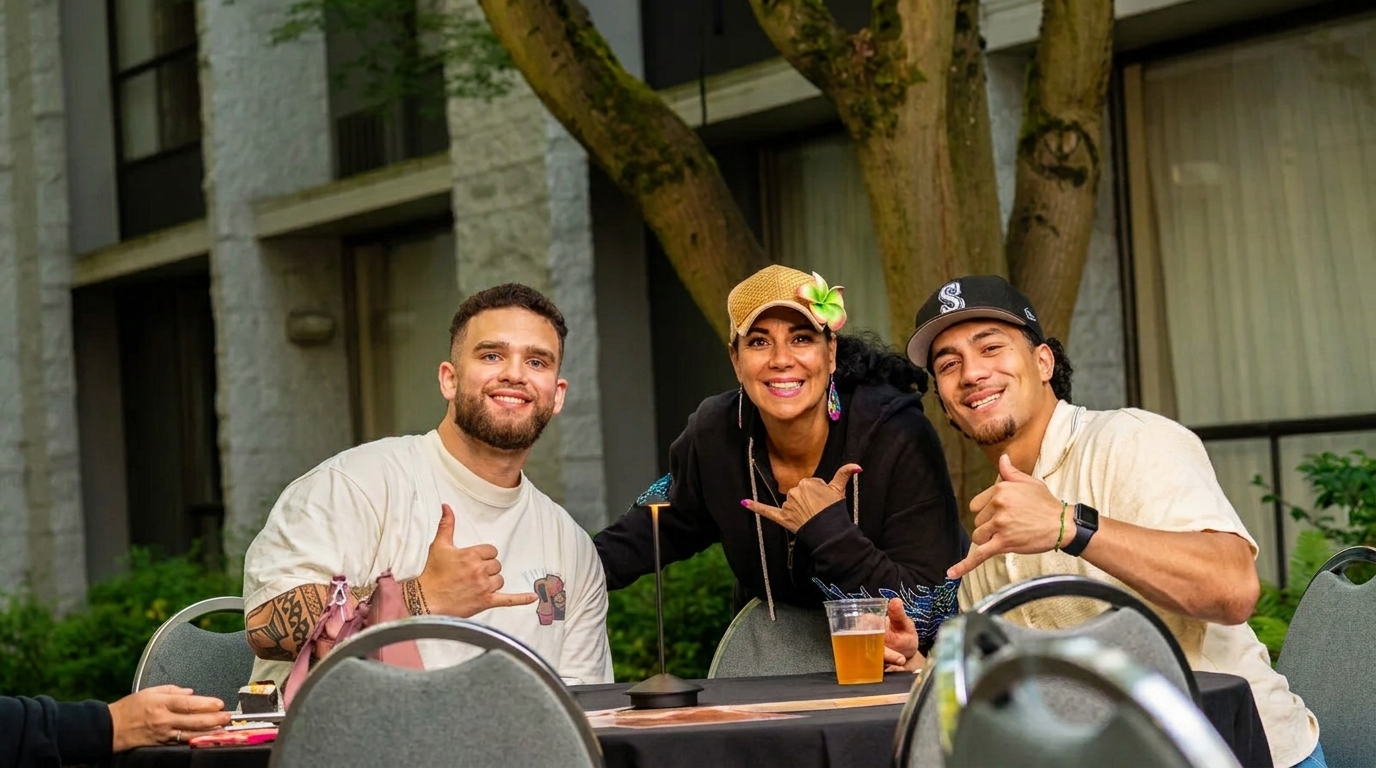 Three smiling people, two men and one woman, giving shaka signs at an outdoor table.