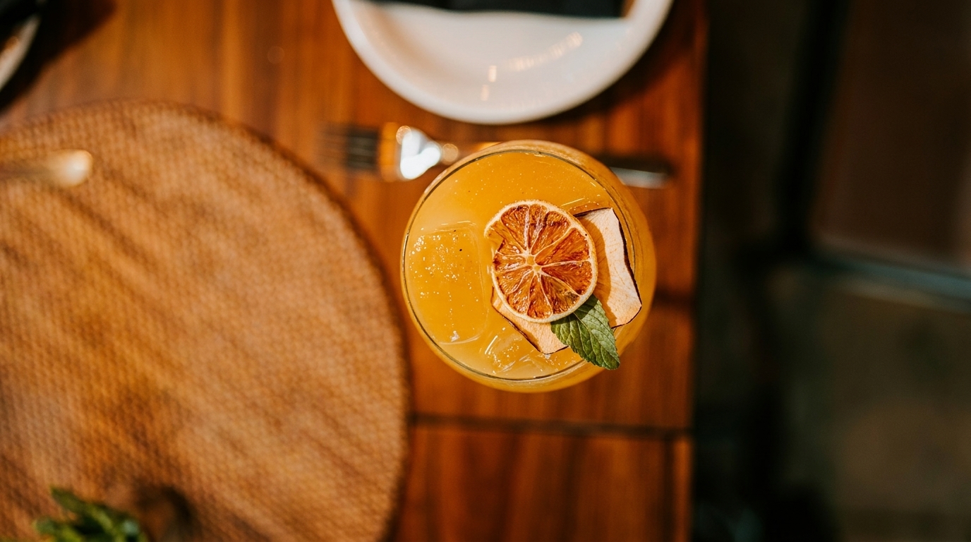 Overhead view of a vibrant orange cocktail garnished with dried citrus and mint on a wooden table.