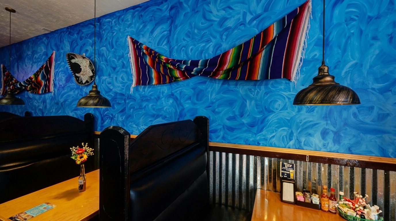 A vibrant blue wall with Mexican blankets and sombreros, black booths, and wooden tables with condiments.