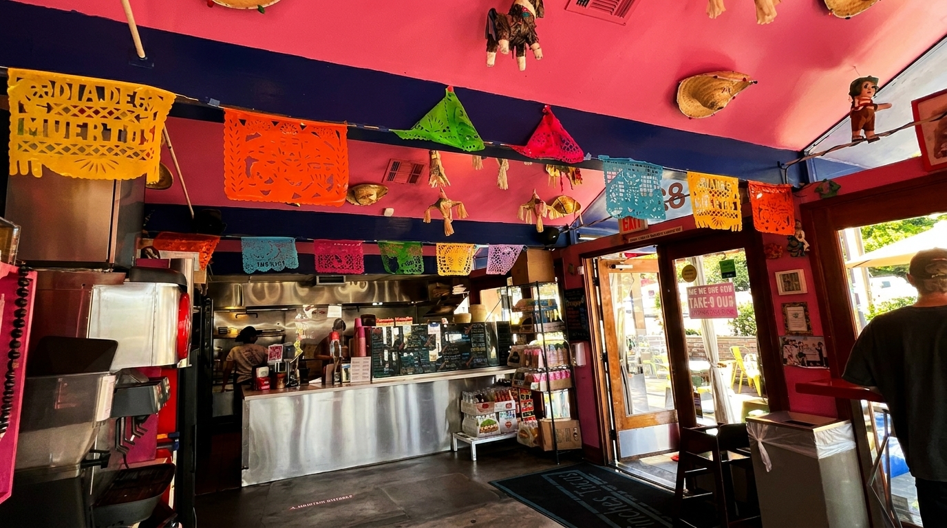 Colorful Mexican papel picado banners adorn a vibrant pink and blue restaurant interior with an open kitchen.