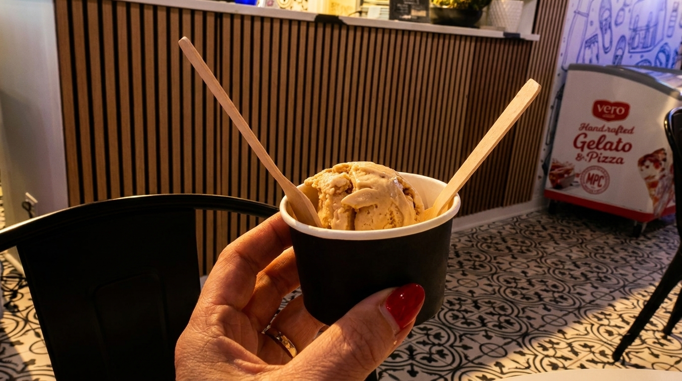 Hand holding a cup of gelato with two wooden spoons, inside a Vero Gelato & Pizza shop.