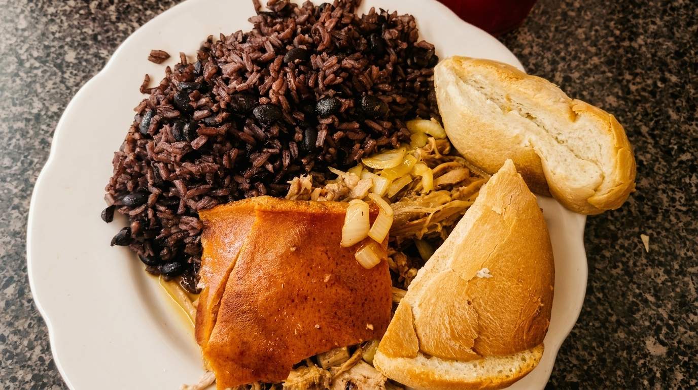 A Cuban meal with black beans and rice, roasted pork with onions, and two pieces of bread.