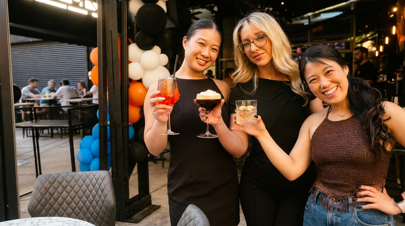 Three smiling women holding cocktails on an outdoor patio with balloons and blurred diners.