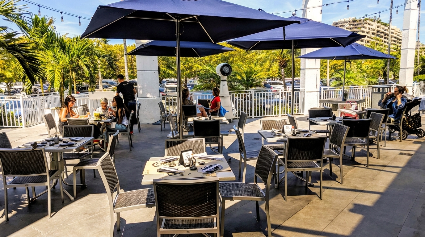 Outdoor restaurant patio with blue umbrellas, dining tables, and patrons enjoying meals under palm trees.