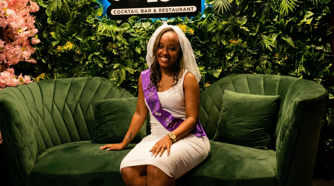 Smiling woman in a white dress, veil, and purple sash sitting on a green velvet couch.