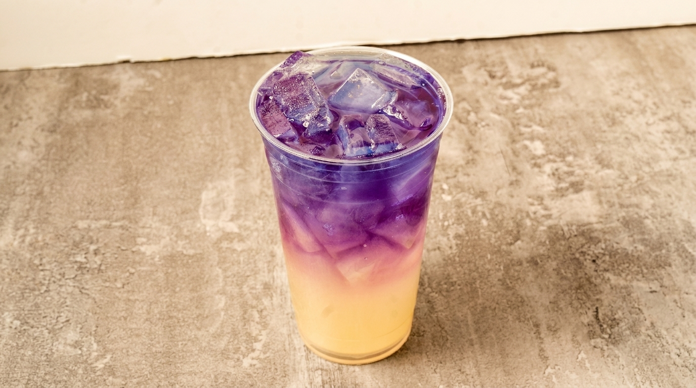 A vibrant purple and yellow layered drink with ice in a clear plastic cup on a textured gray surface.