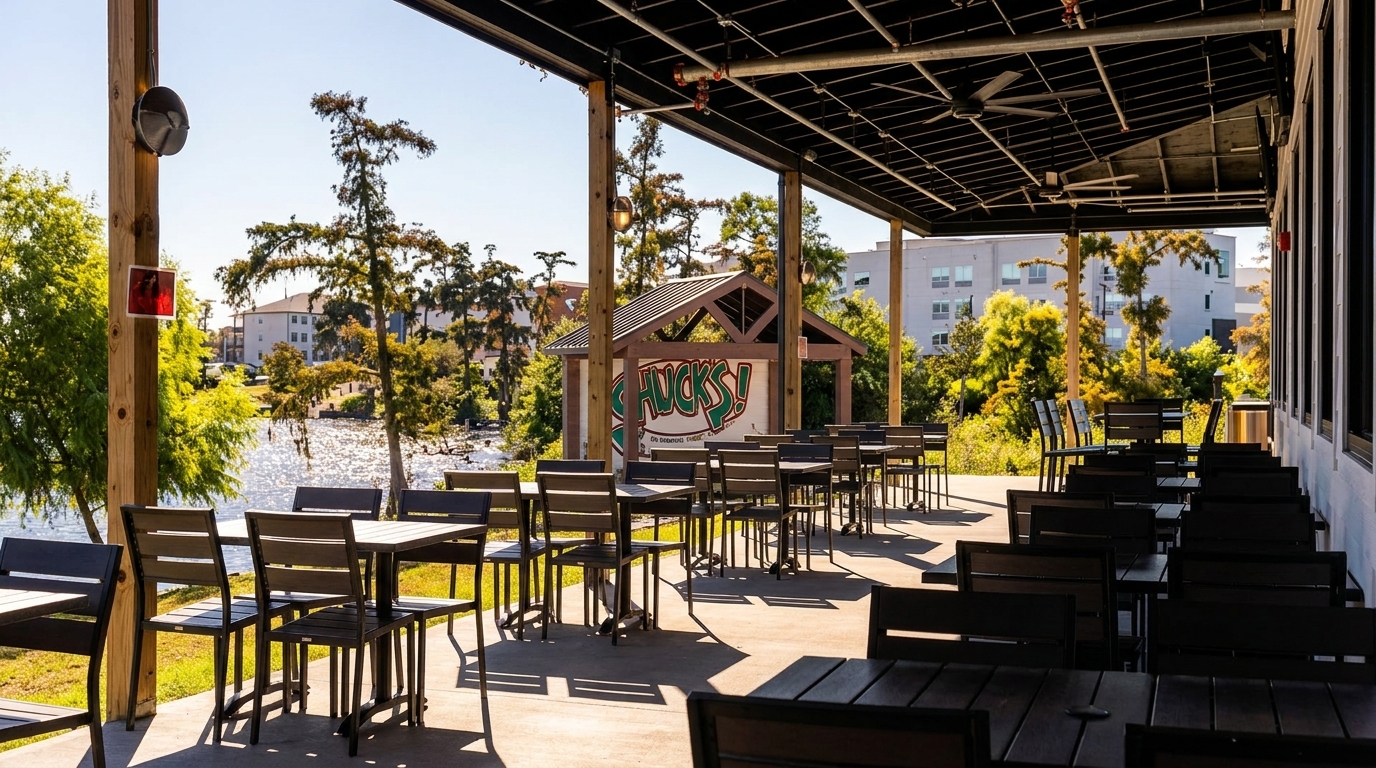 An outdoor patio with empty tables and chairs overlooks a sunny lake and distant buildings.