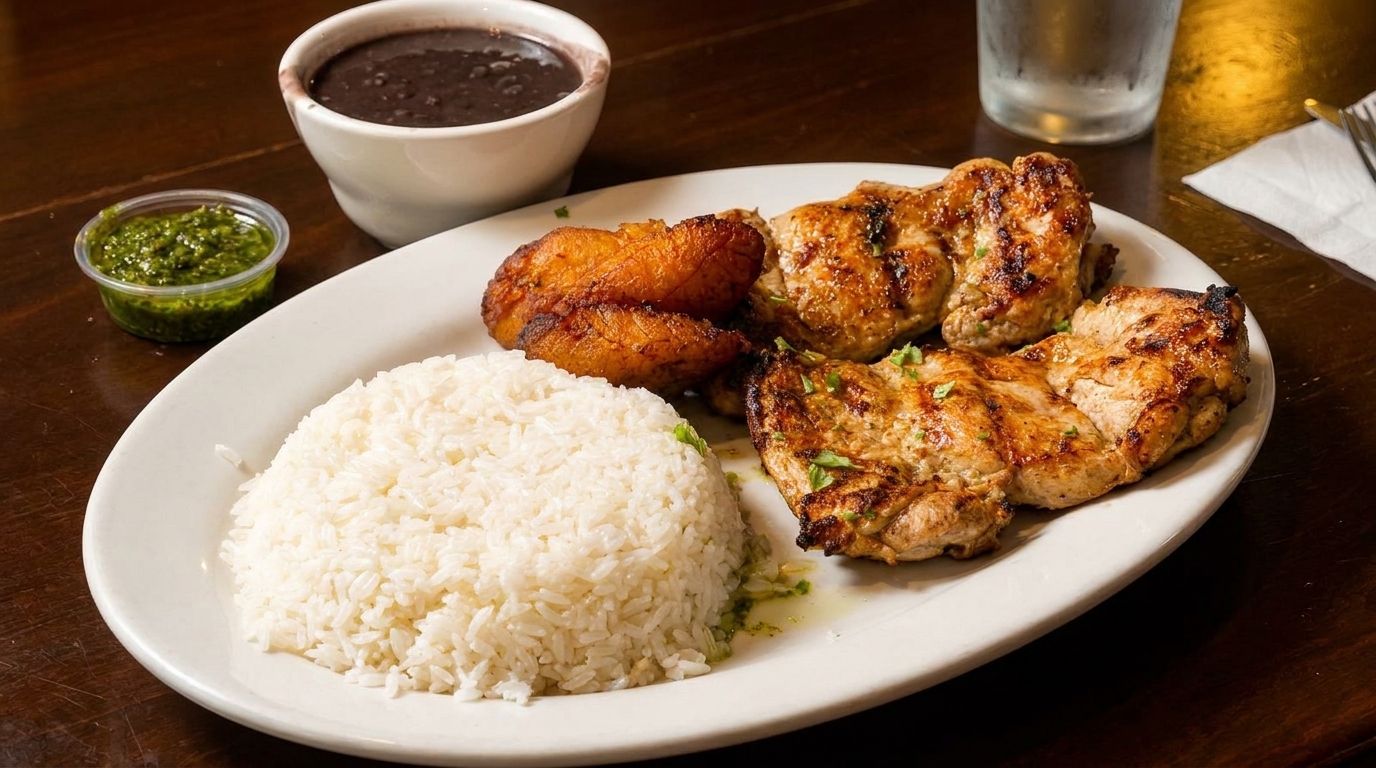 Grilled chicken with white rice, fried plantains, black beans, and green sauce on a white plate.