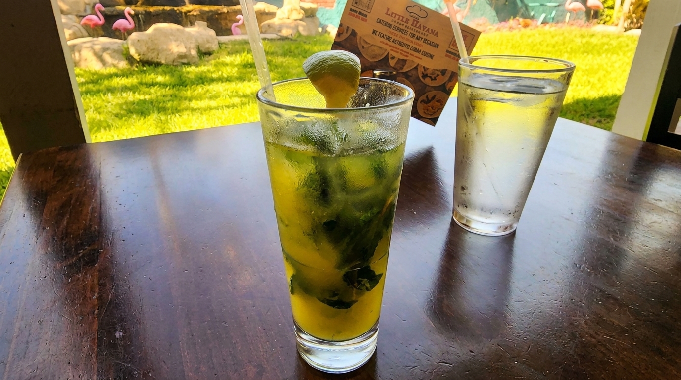 Mojito and water on a dark wooden table with a Little Havana menu and pink flamingos in the background.