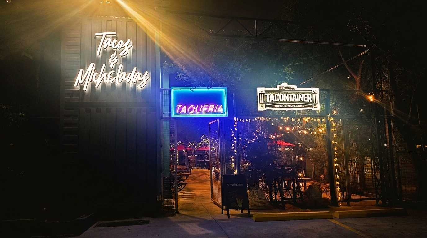Exterior of "Tacos & Micheladas" restaurant at night, with neon signs and string lights illuminating the entrance.