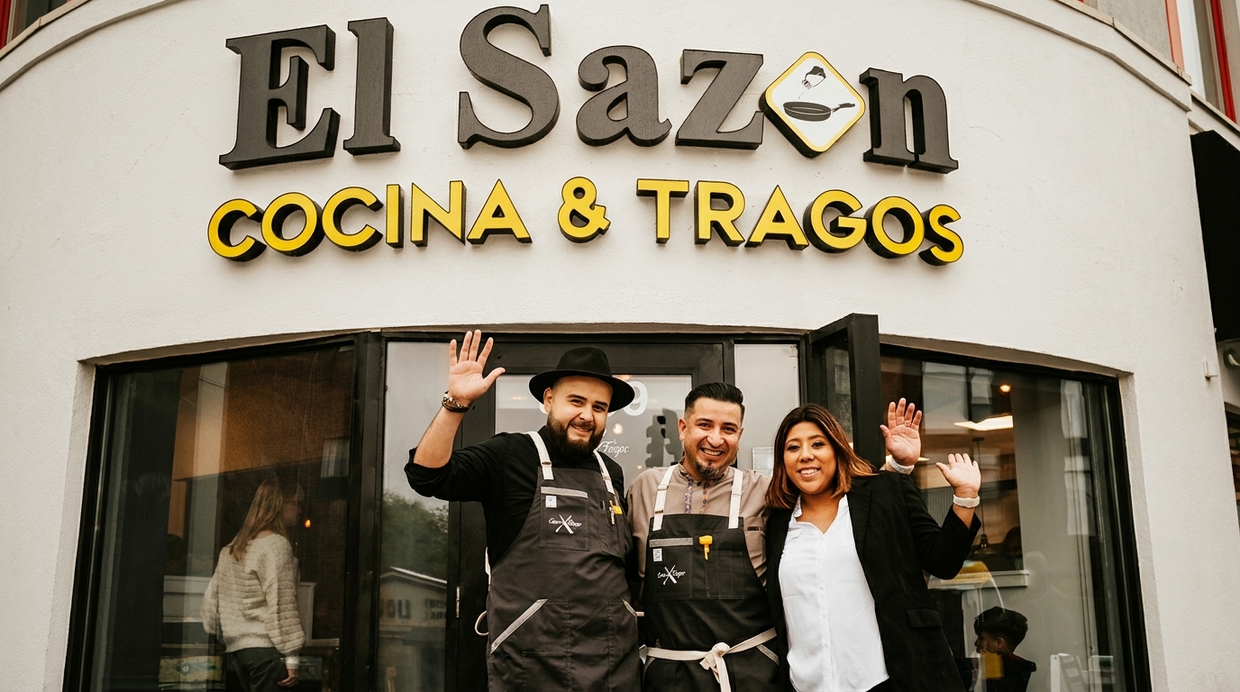 Three smiling people, two men and one woman, wave in front of the El Sazon restaurant sign.