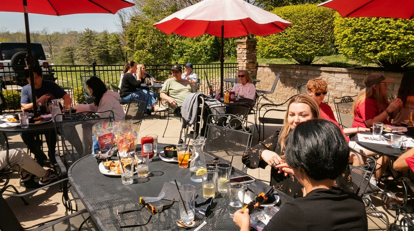 Patrons enjoy food and drinks on a sunny outdoor patio with red umbrellas and lush greenery.