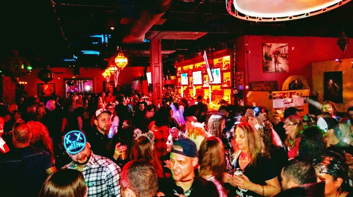A crowded bar scene with people socializing, some wearing costumes, under dim red and blue lighting.