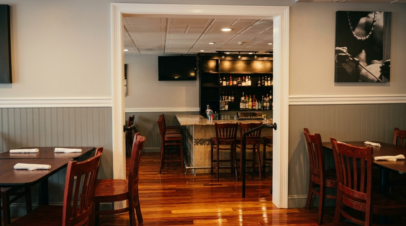 A restaurant dining room with wooden tables and chairs, leading to a bar area with liquor bottles.