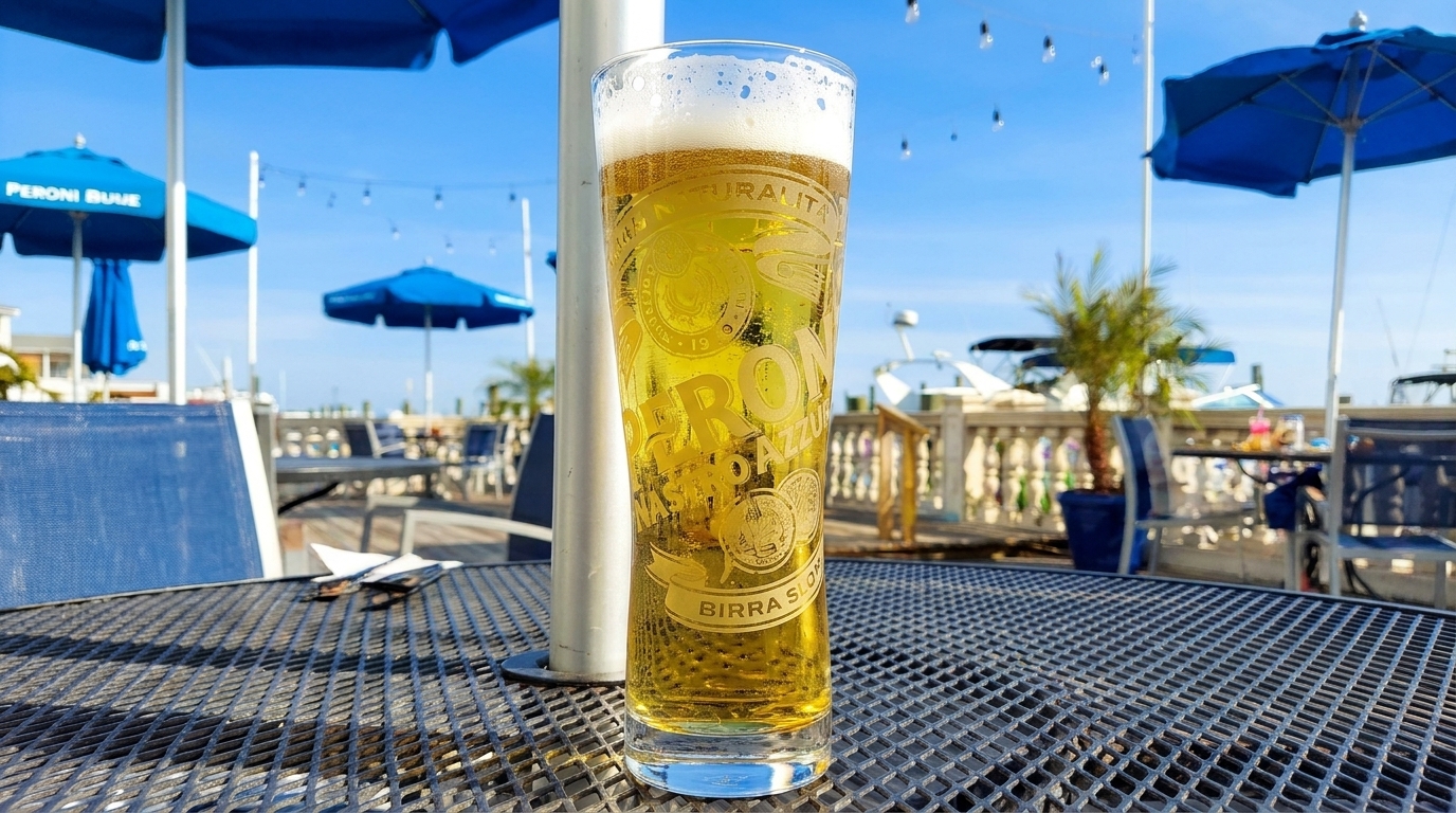 A glass of Peroni beer on a metal table at an outdoor restaurant with blue umbrellas and boats.