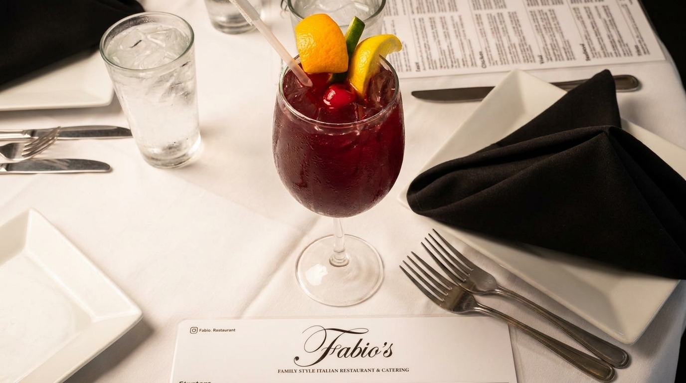 A red cocktail with fruit garnishes and a water glass on a white tablecloth at Fabio's restaurant.