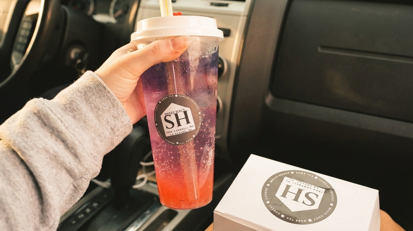 Hand holding a Sweet Haus boba tea with a box of treats in a car.