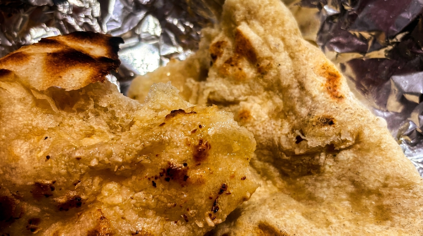 Close-up of warm, golden-brown naan bread with charred spots, resting on crinkled aluminum foil.