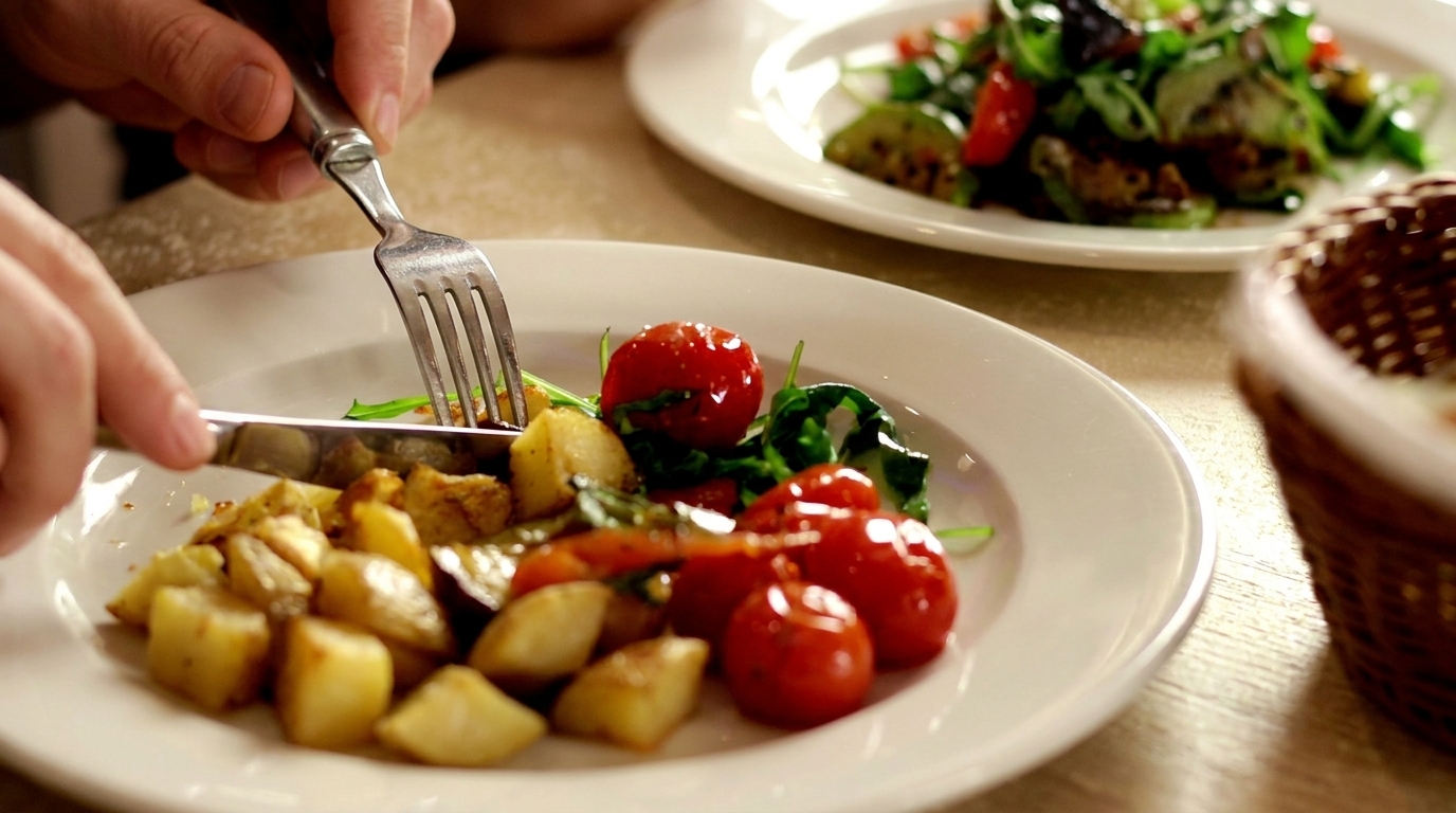 Hands cutting roasted potatoes and cherry tomatoes on a white plate with a salad in the background.