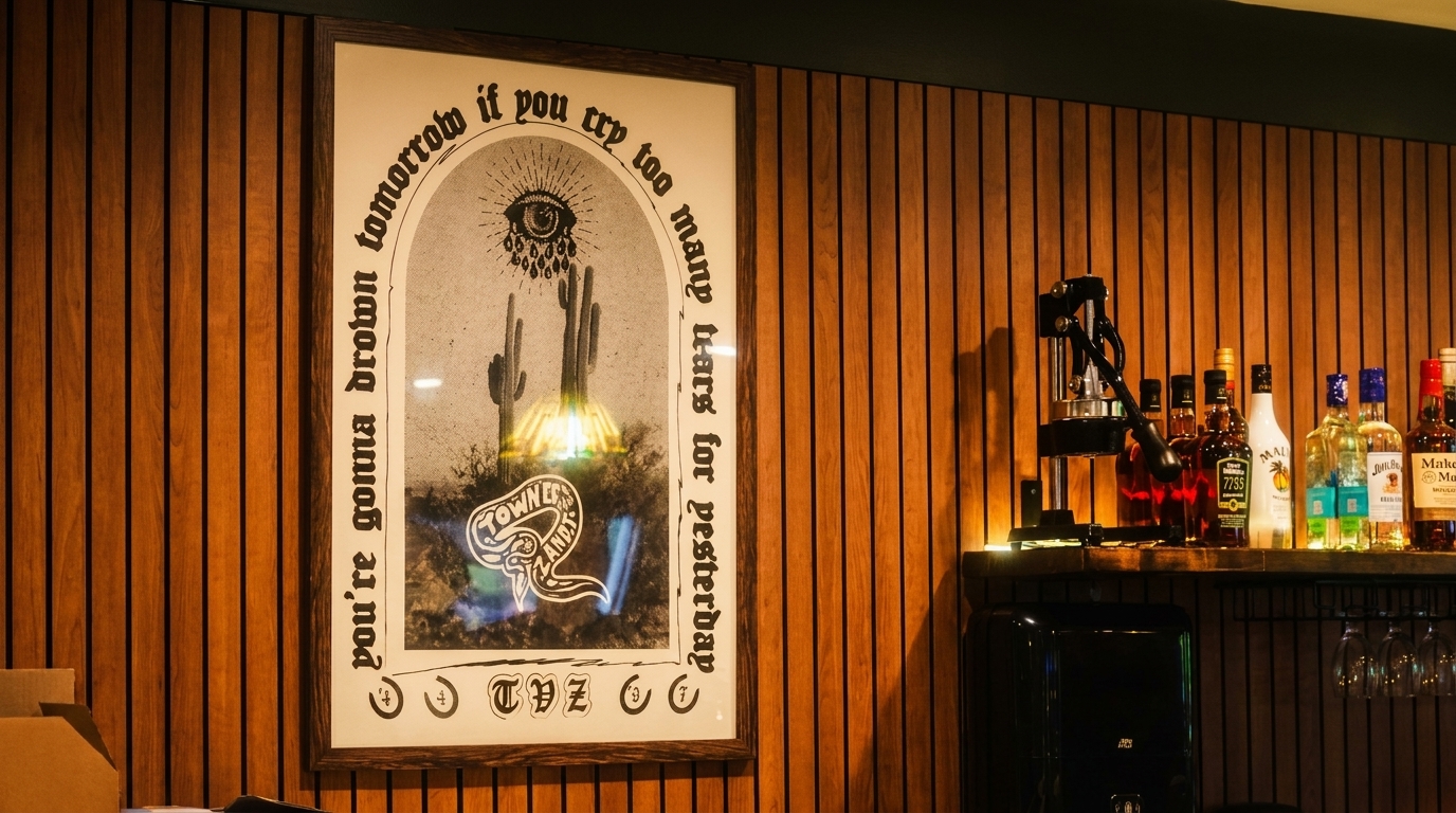 A framed poster with an eye, cacti, and text hangs on a wooden slatted wall next to a bar shelf.