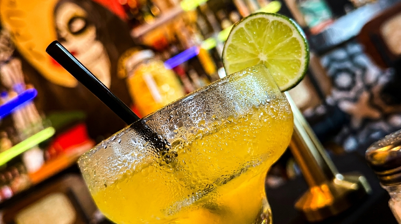 A vibrant yellow margarita with a lime wedge and black straw, condensation on the glass.