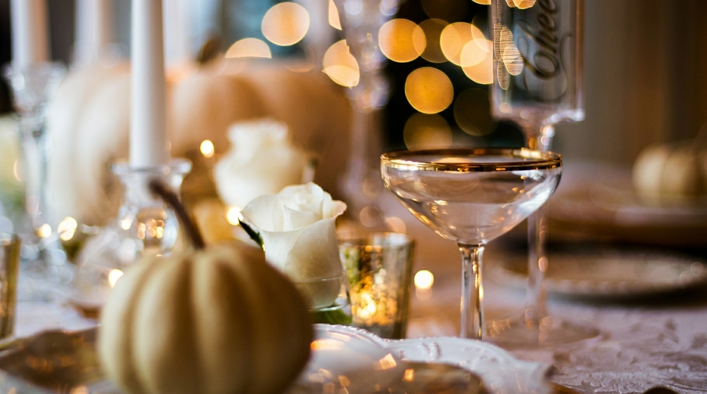 Elegant table setting with white pumpkins, roses, candles, and gold-rimmed glasses, with bokeh lights.