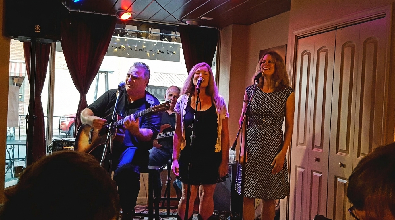 A band performs live, with a man playing guitar and two women singing into microphones.