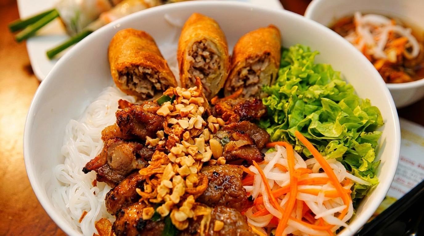 A close-up of a Vietnamese noodle bowl with spring rolls, grilled meat, peanuts, and fresh vegetables.