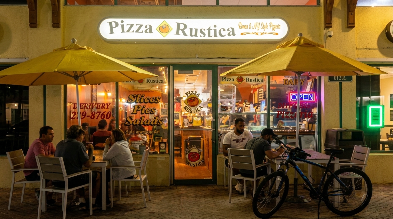 Pizza Rustica storefront at night with outdoor seating, customers, and a bicycle parked nearby.