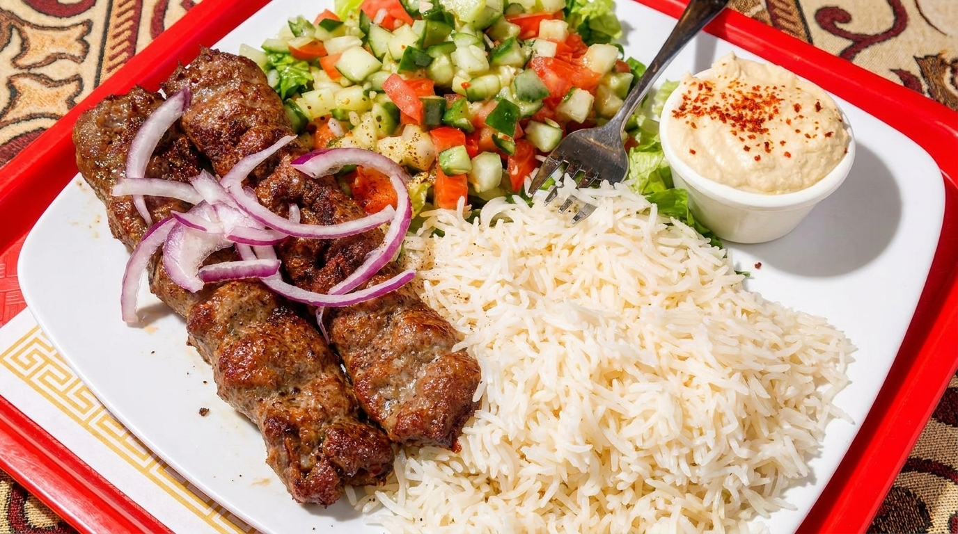 Grilled meat skewers with red onion, rice, chopped salad, and hummus on a white plate.
