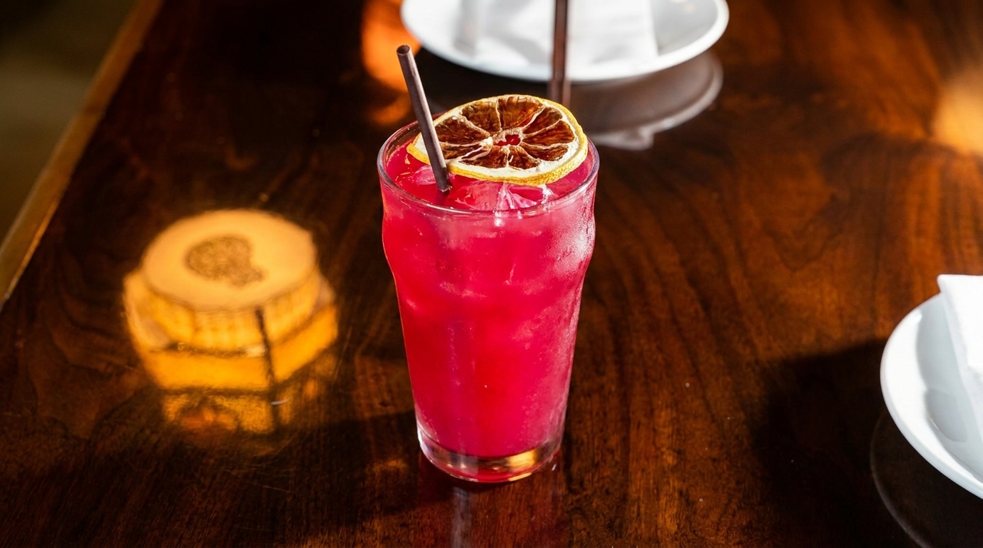 Vibrant red cocktail with ice, a straw, and a dried orange slice on a dark wooden table.