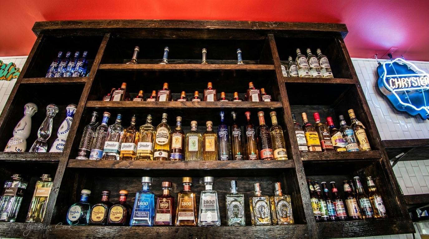 A rustic wooden shelf filled with various bottles of tequila and other spirits, with a neon sign in the background.