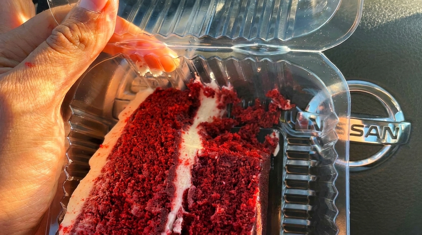 Hand holding a plastic container with a partially eaten slice of red velvet cake, with a car logo in the background.