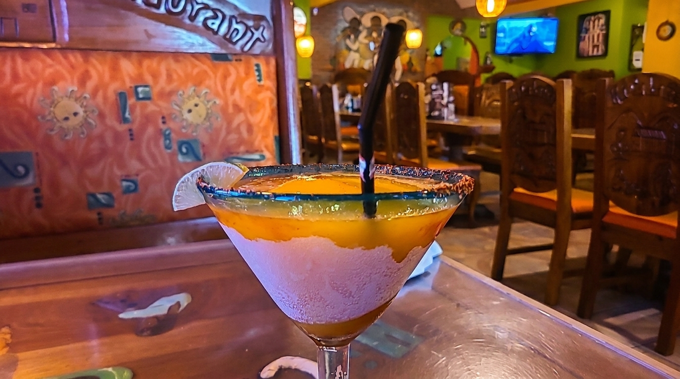 A colorful layered margarita with a lime wedge and straw sits on a table in a vibrant restaurant.
