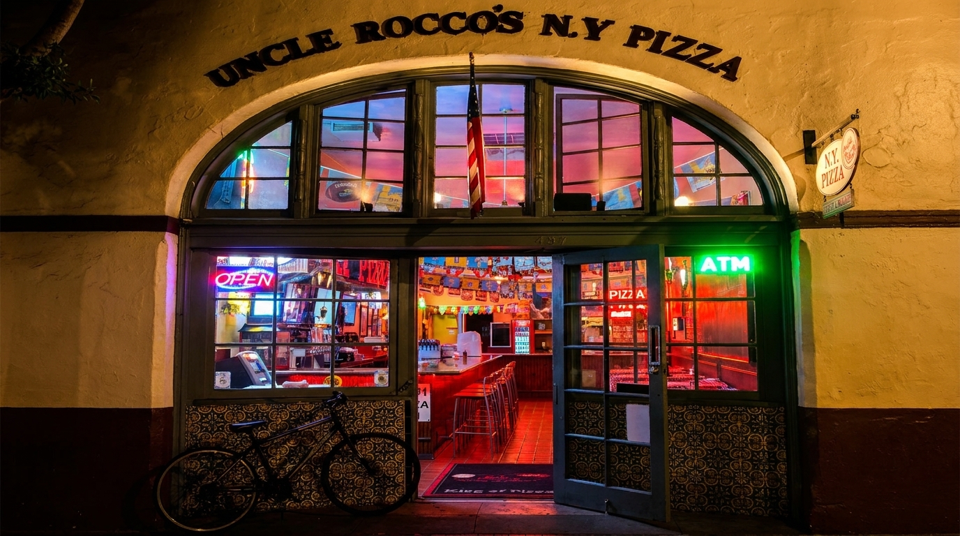 Uncle Rocco's N.Y. Pizza storefront at night, with neon signs, a bicycle, and an open door.