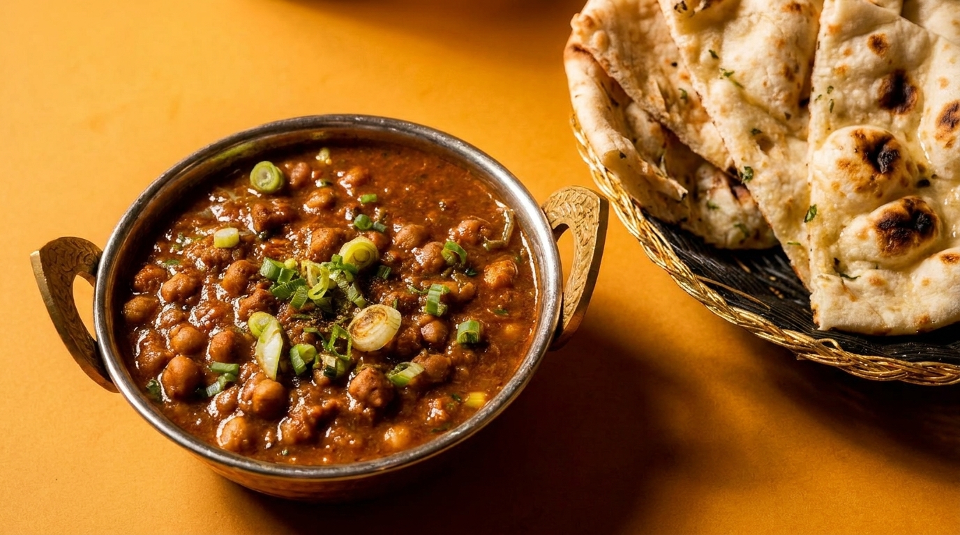 A metal bowl of chana masala with green onions next to a basket of naan bread.