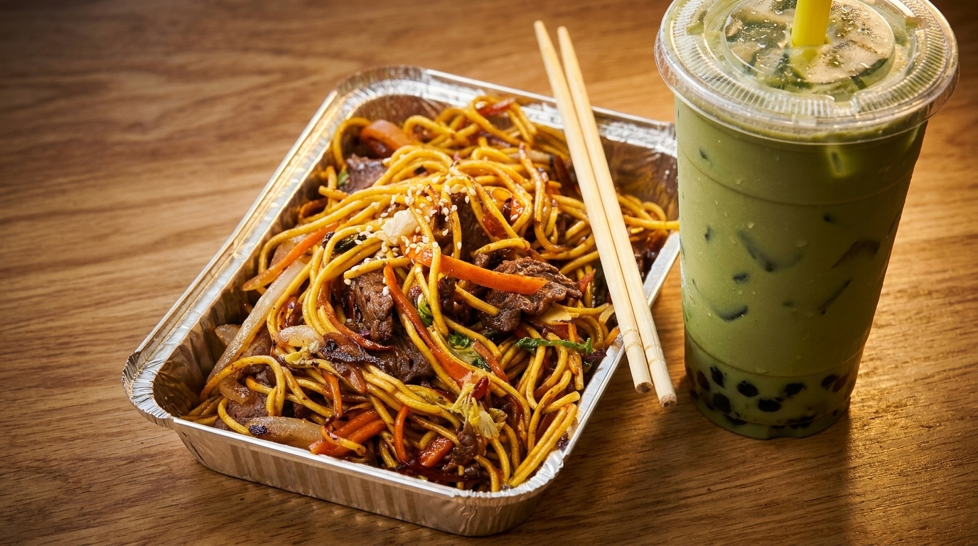 Beef chow mein in a foil container with chopsticks and a green boba tea on a wooden table.