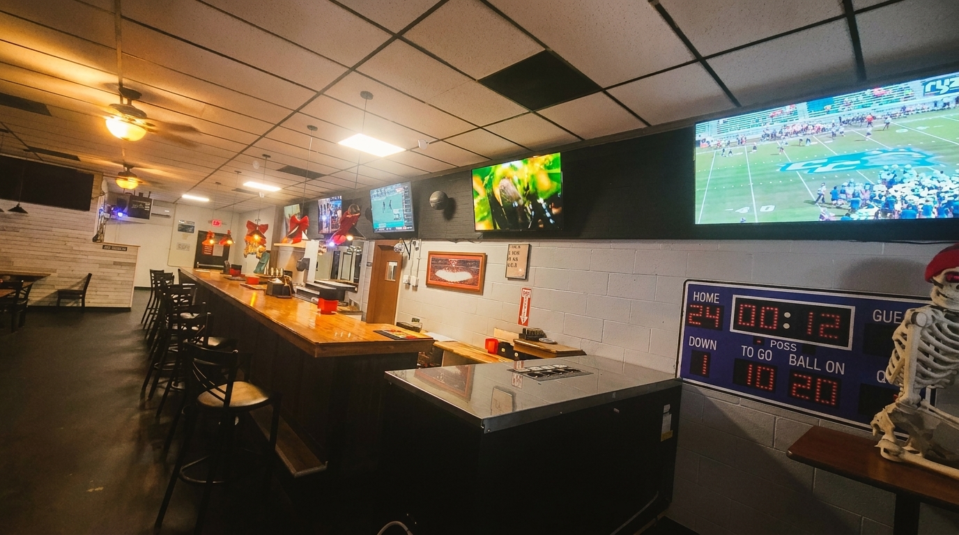 A dimly lit bar with a long wooden counter, bar stools, and multiple TVs showing sports.