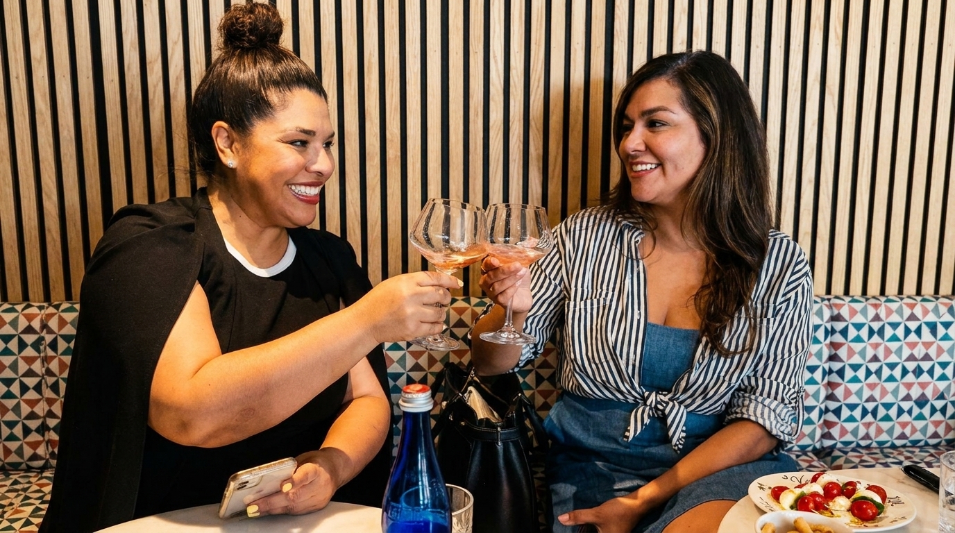 Two women smiling, toasting with glasses of rosé wine in a restaurant booth.