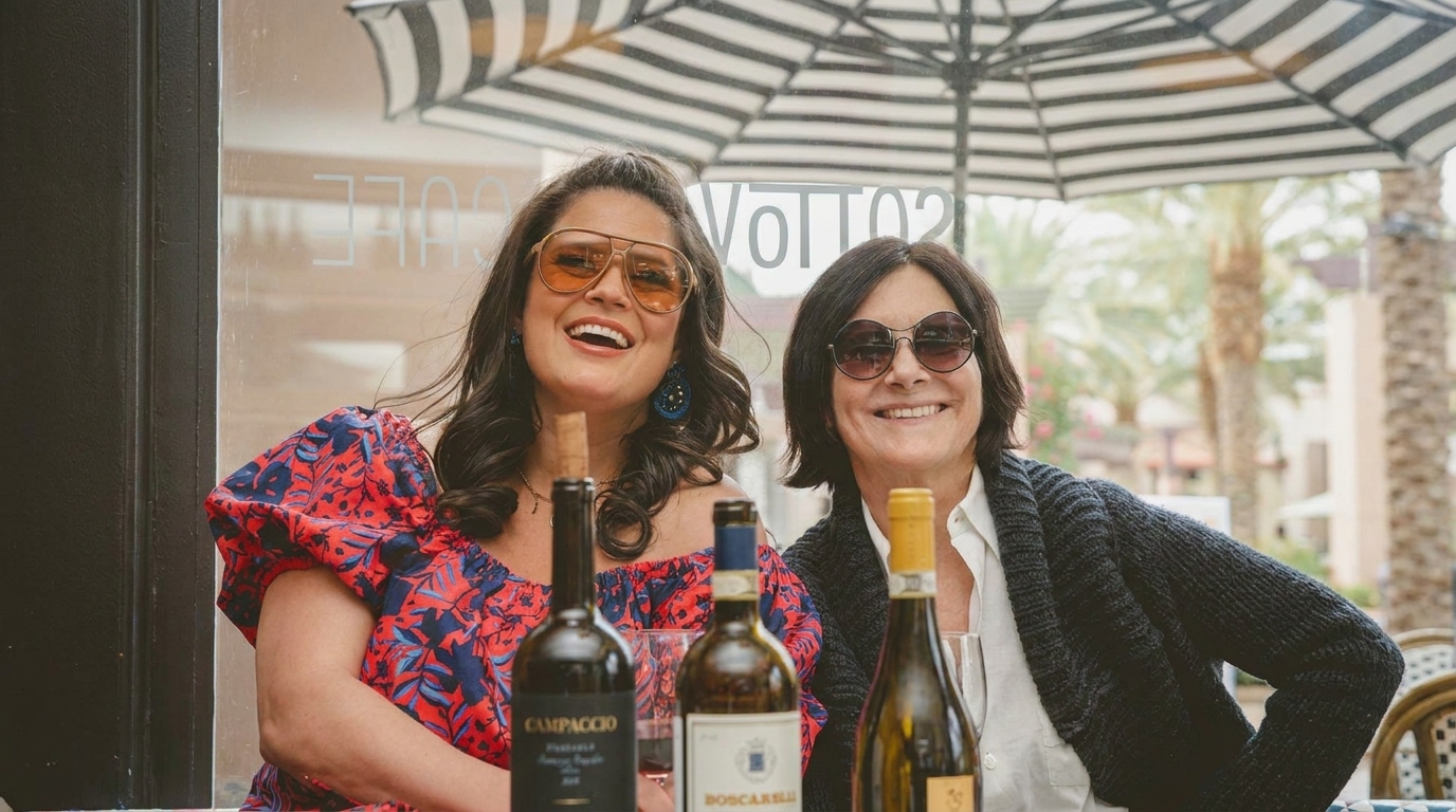 Two smiling women in sunglasses sit at an outdoor cafe table with wine bottles.