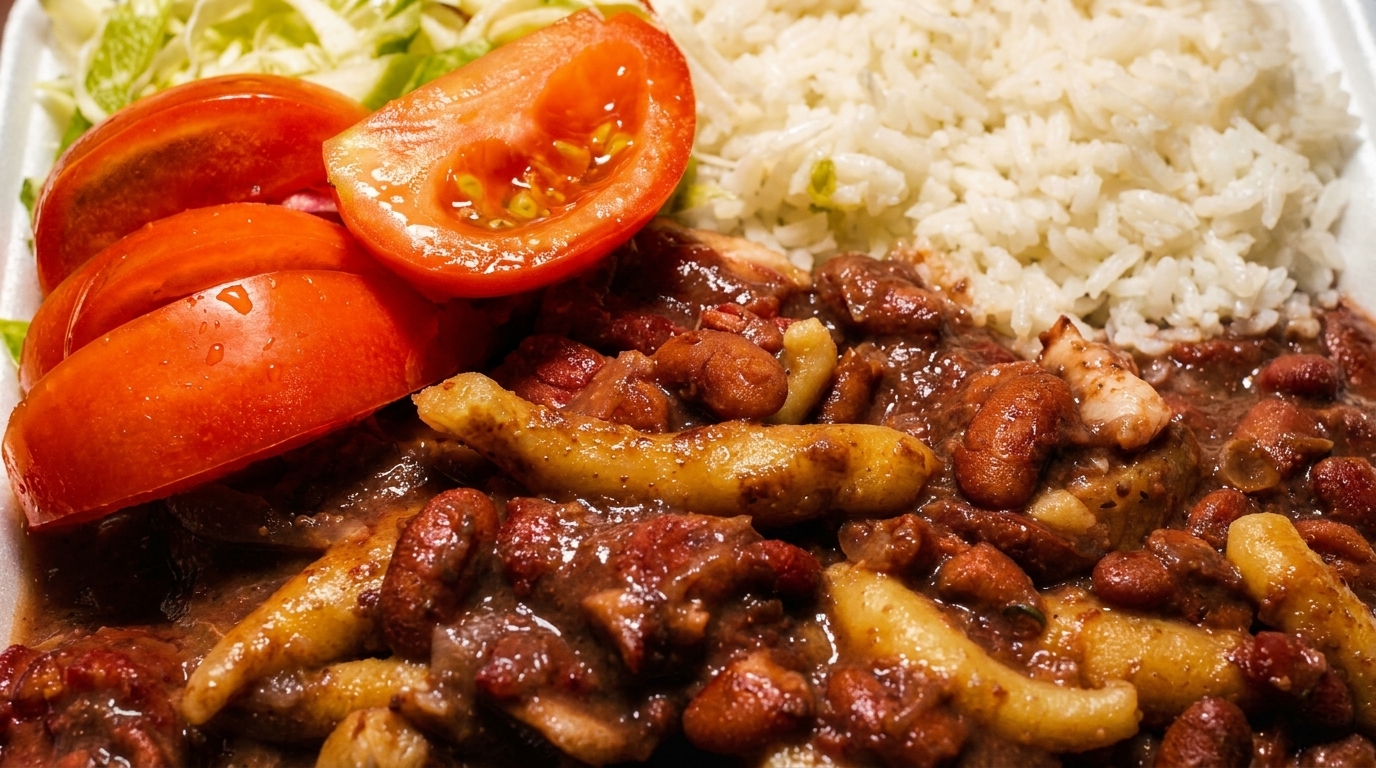 Close-up of a Jamaican meal with stewed chicken and beans, rice, sliced tomatoes, and shredded lettuce.