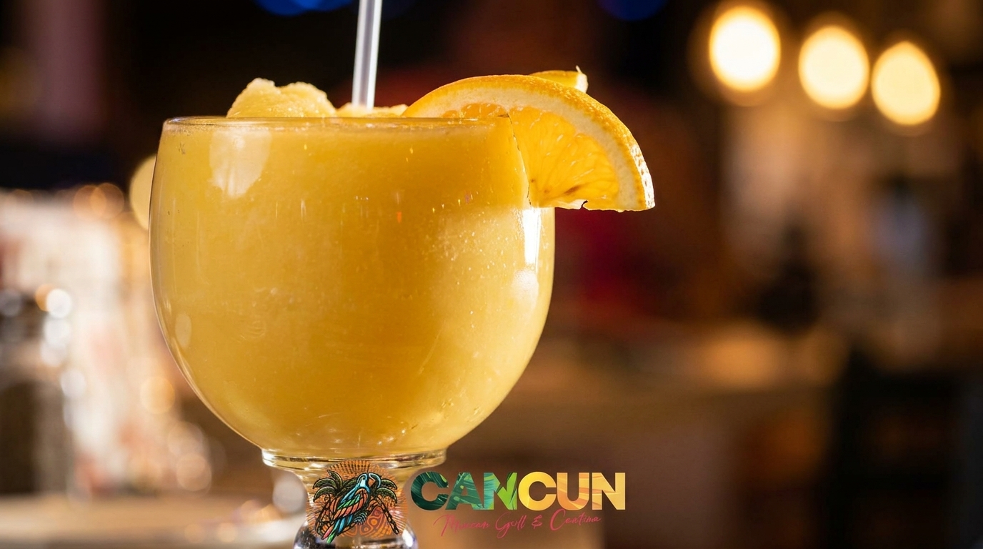 A vibrant yellow frozen drink, garnished with an orange slice, in a Cancun Mexican Grill & Cantina glass.