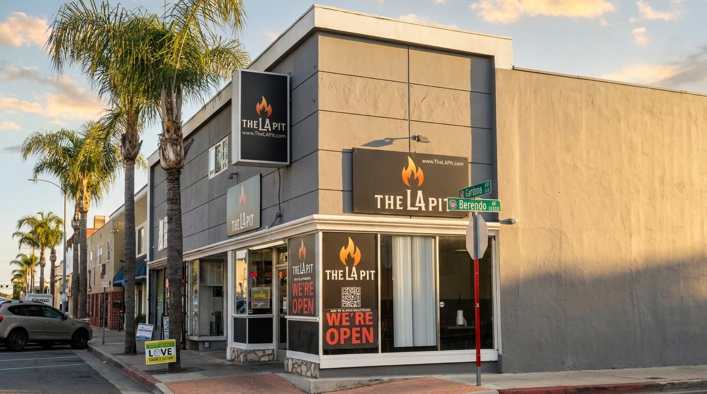 The LA Pit restaurant exterior with "We're Open" signs, palm trees, and a street sign at sunset.