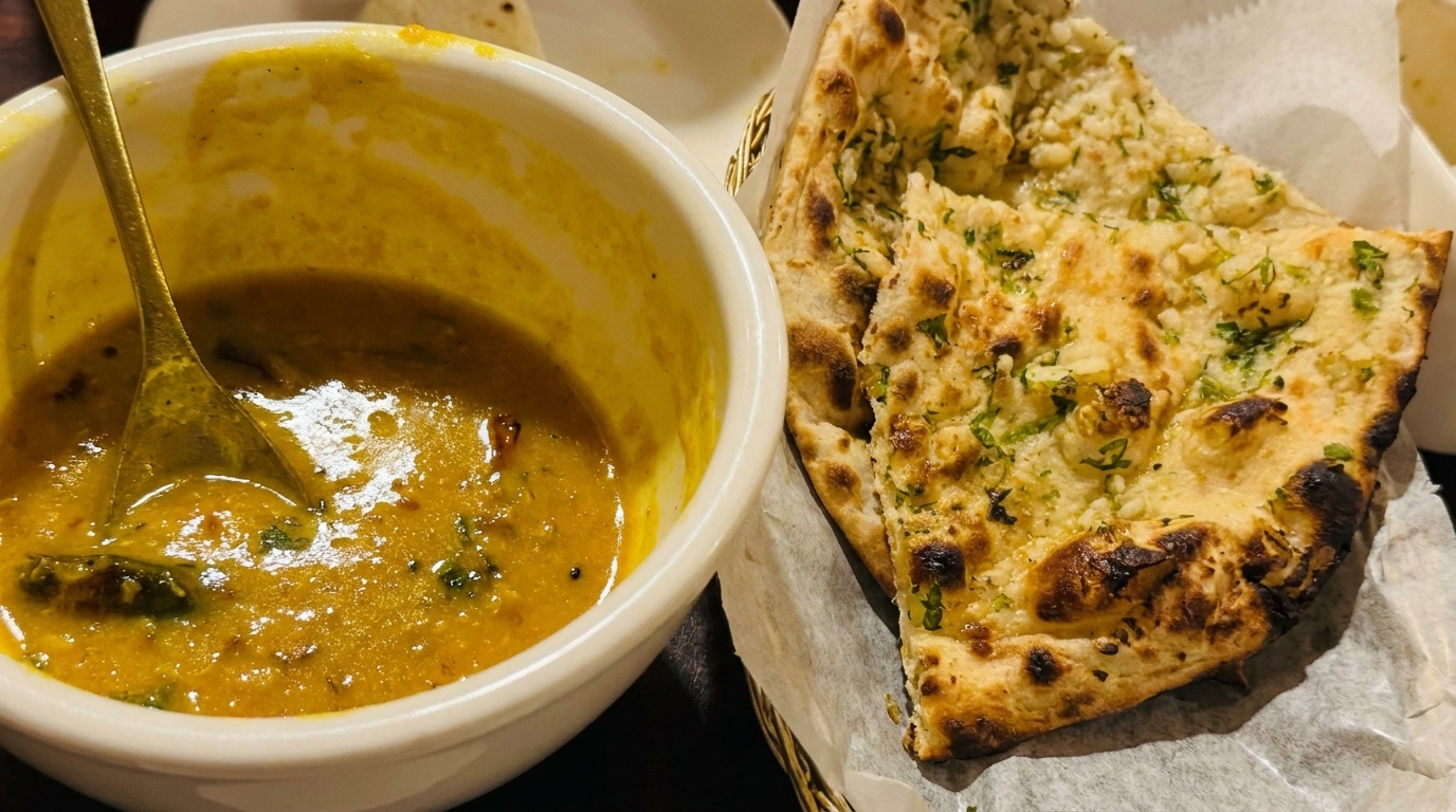 A bowl of yellow lentil dal with a spoon next to a basket of garlic naan bread.